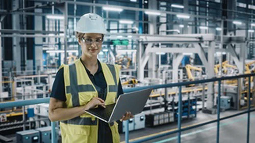 A woman wearing a hardhat and holding a laptop in a factory.