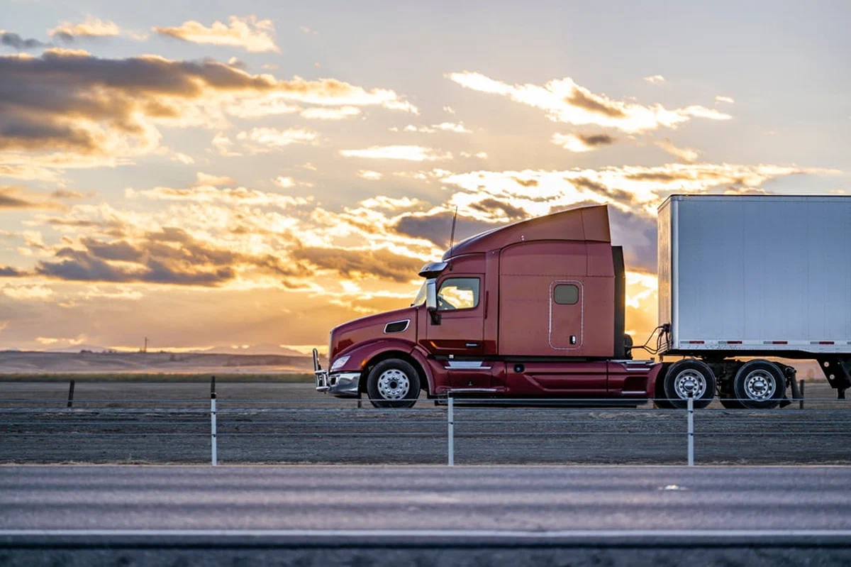 A photograph of a red semi truck driving on a road with mountains in the background.