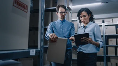 A man and a woman standing in a stock room with boxes marked as fragile surrounding them.