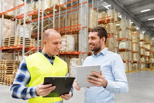 Two men standing in a warehouse looking at clipboards.
